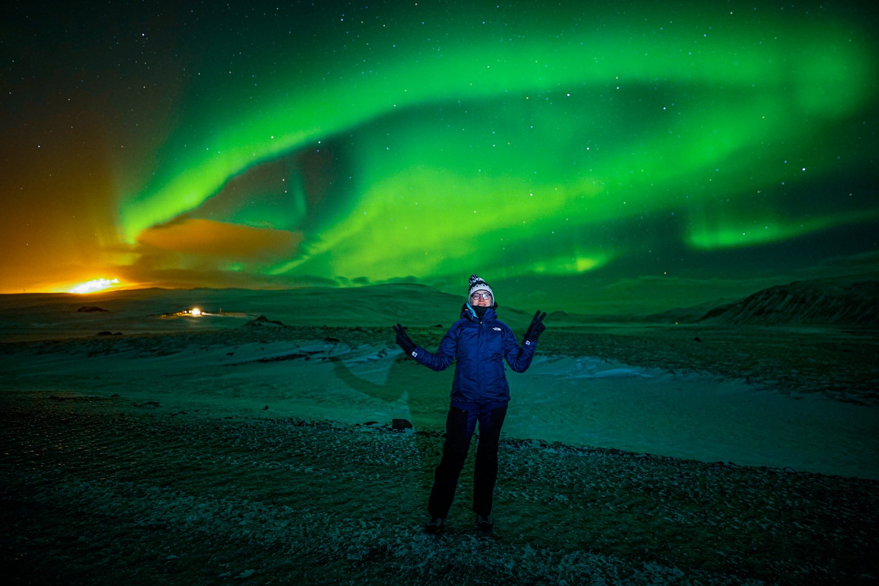 A guest with arms raised under a wide green aurora above a frozen lake