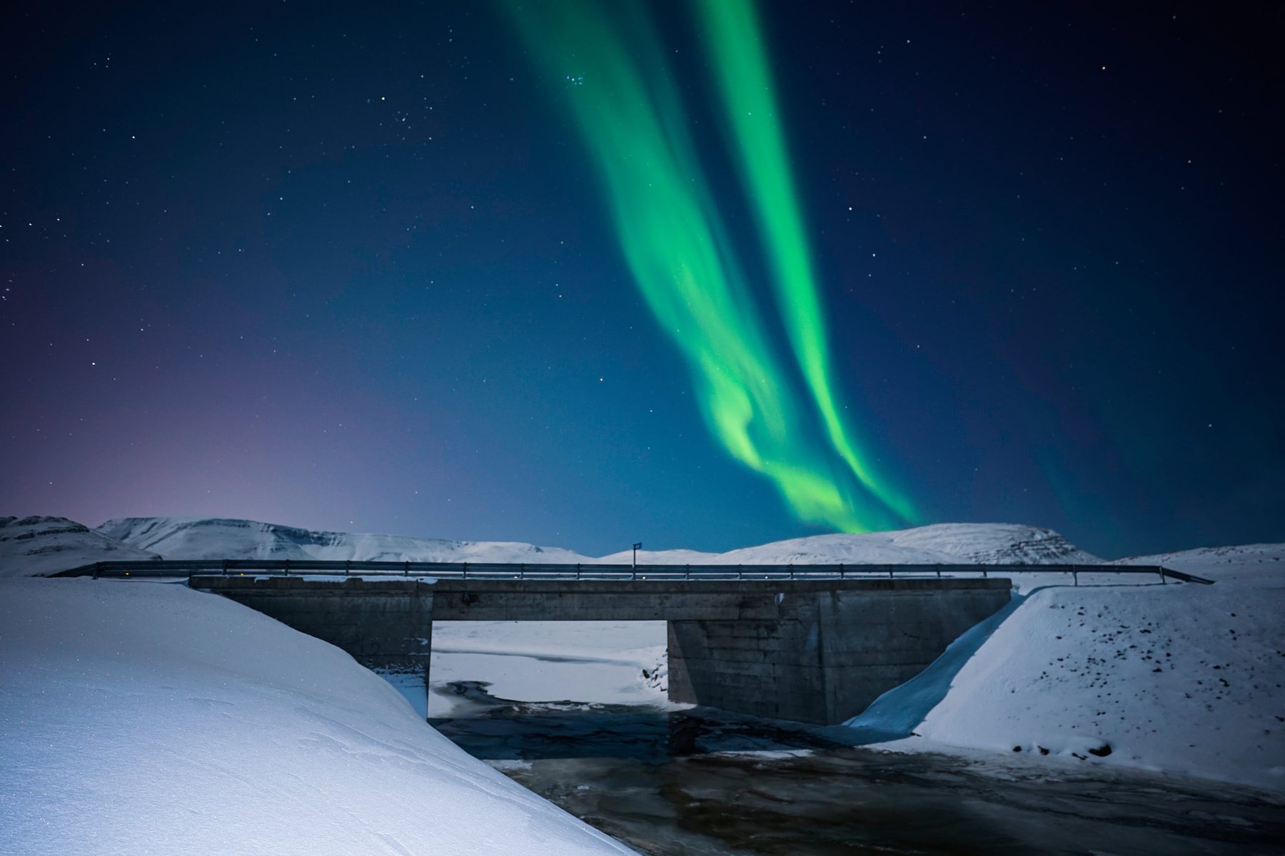 Aurora curtains above a snow covered bridge and mountain ridge