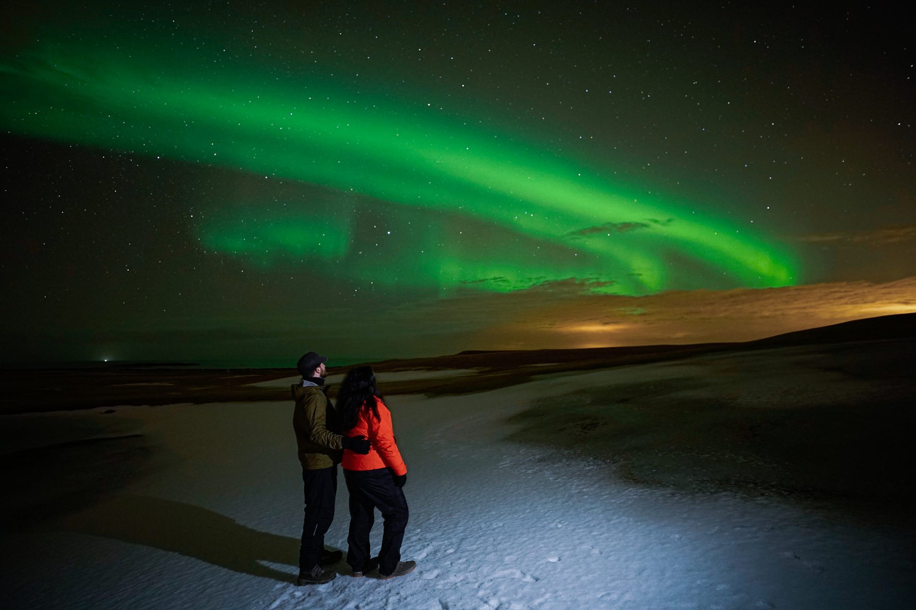 A couple standing together on ice while aurora curls across the sky behind them