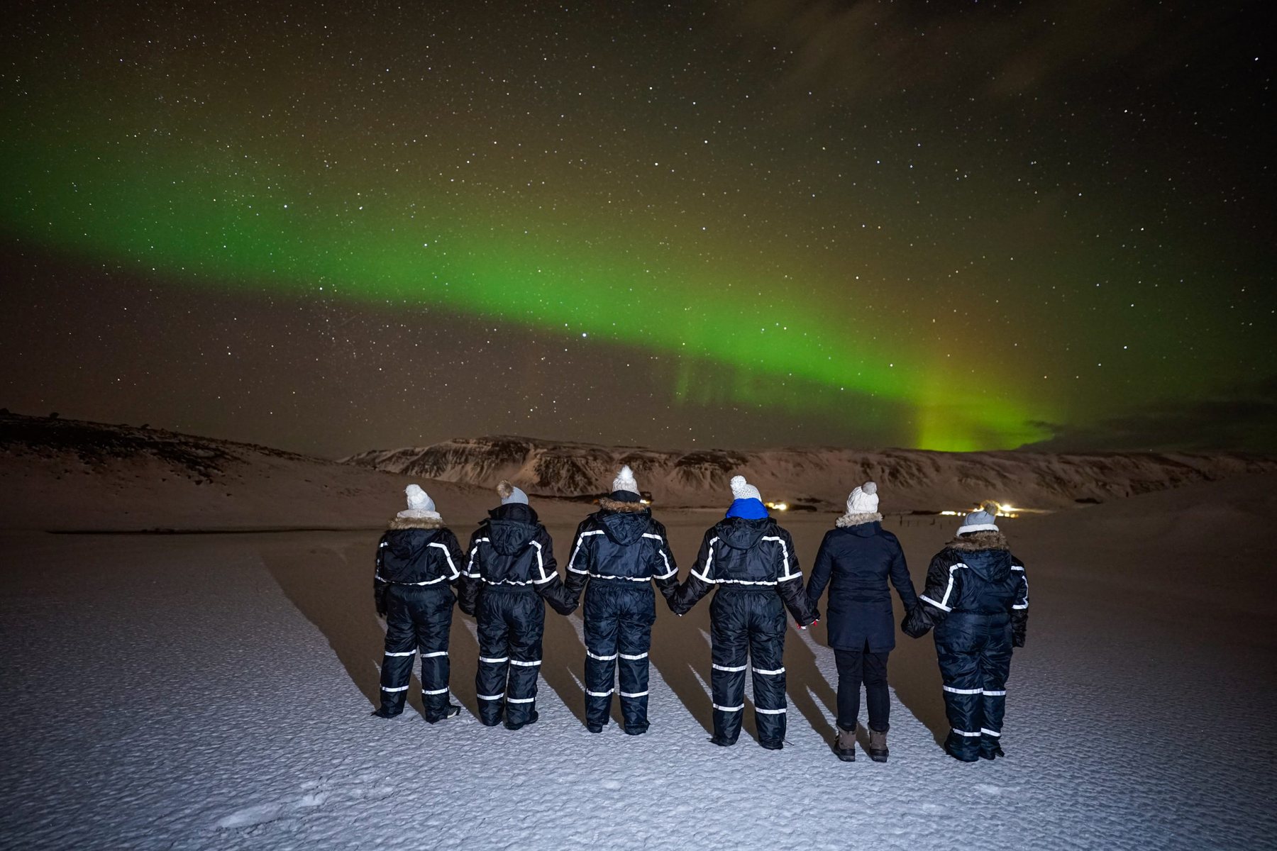 Guests in winter jumpsuits watching the northern lights from outdoor chairs