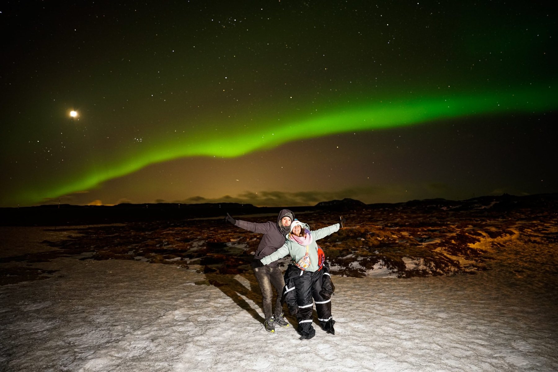 Two guests pointing up at a long green aurora band over a lava field