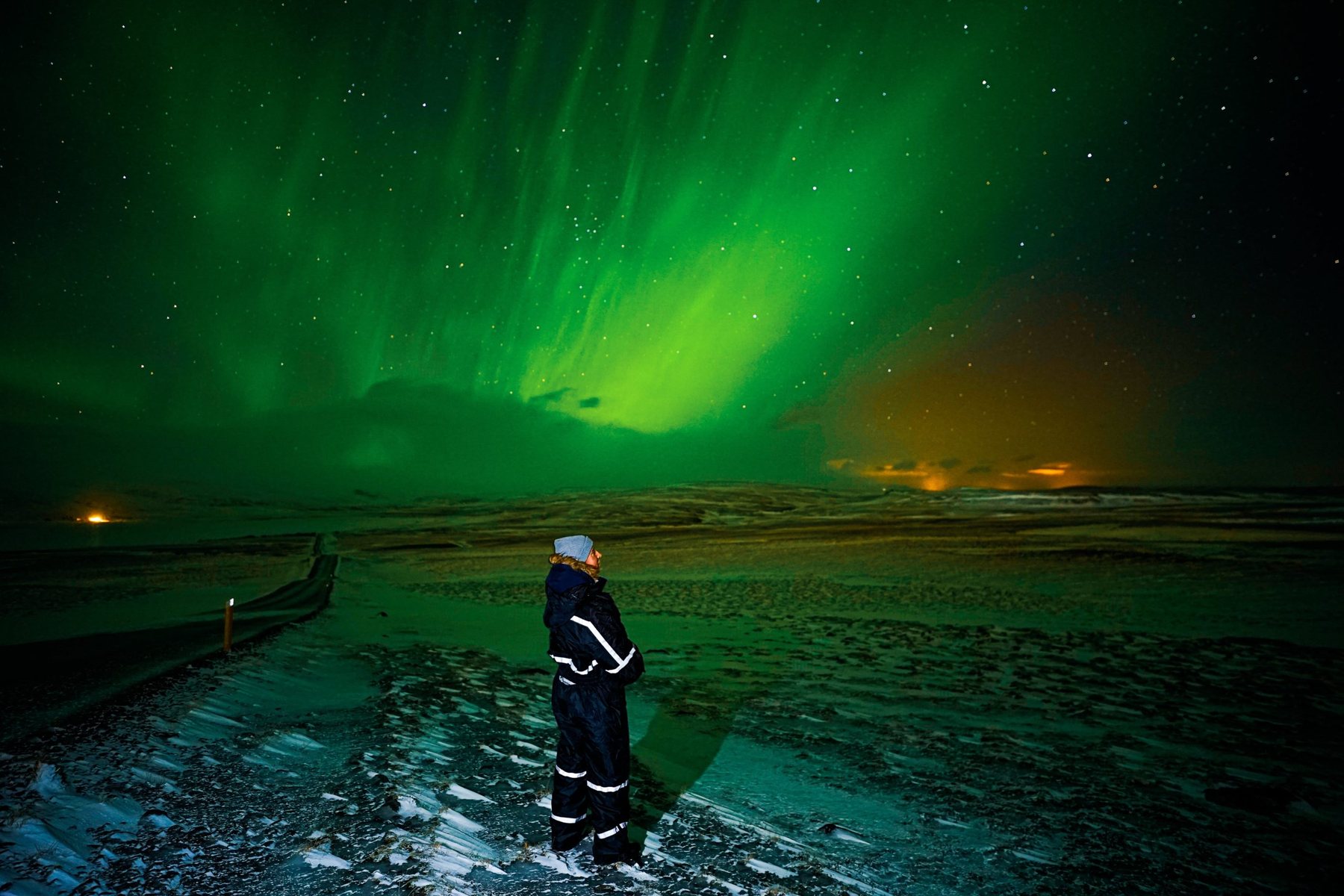 A guest in a thermal jumpsuit standing on frozen ground under bright aurora