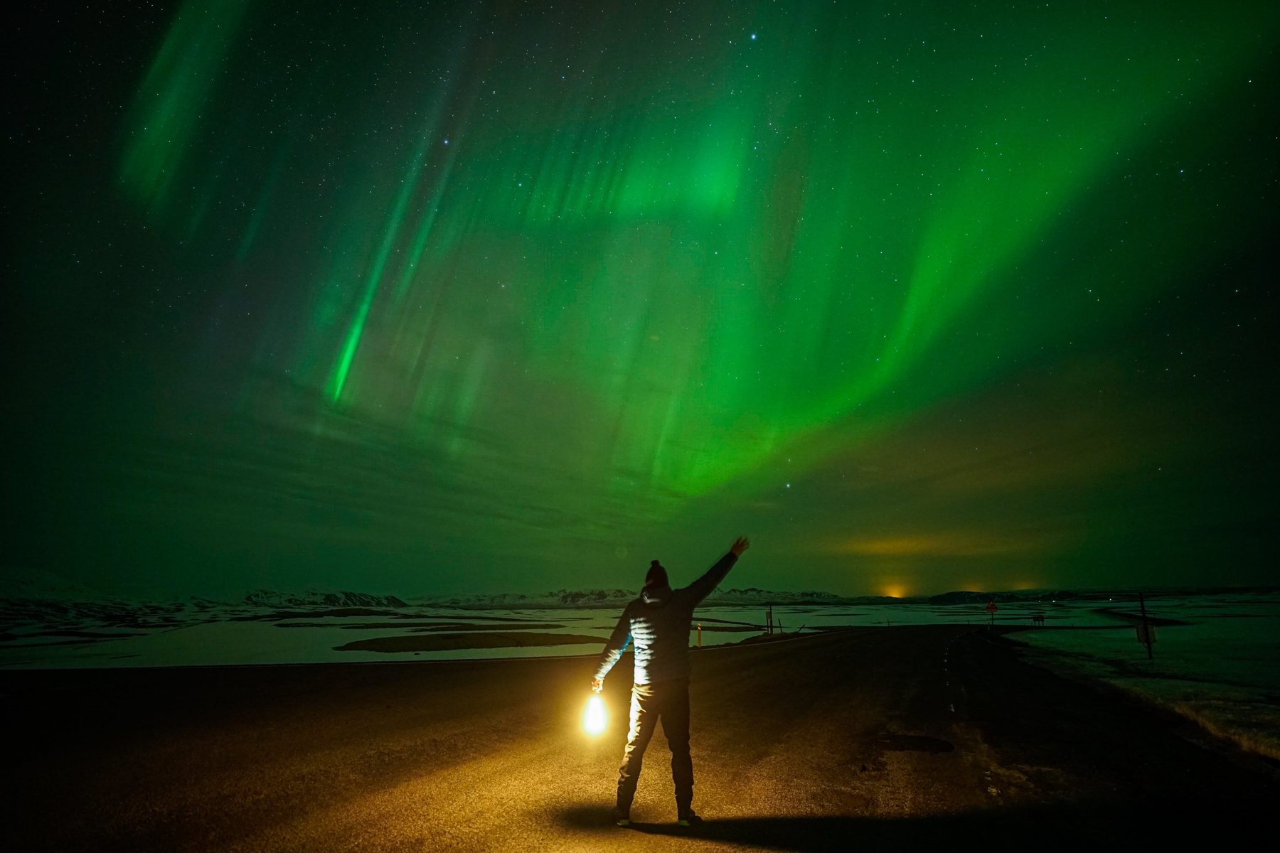 A guest holding a lantern on a dark road with vertical aurora rays above